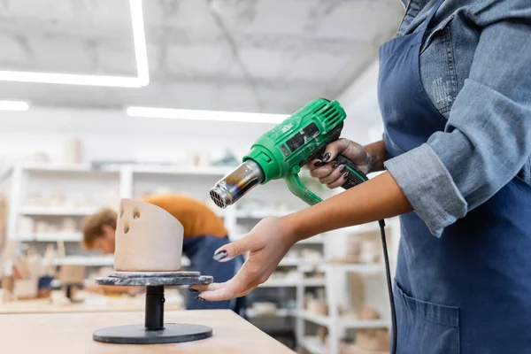 Vue recadrée de femme afro-américaine en tablier séchage sculpture en argile avec pistolet à chaleur dans l'atelier de poterie — Photo de stock