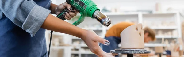 Vue recadrée d'un artisan afro-américain desséchant une sculpture en céramique avec pistolet à chaleur dans un atelier de poterie, bannière — Photo de stock