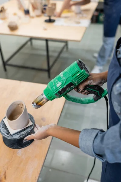 Vista recortada del producto de cerámica de secado artesanal afroamericano con pistola de calor en el taller - foto de stock