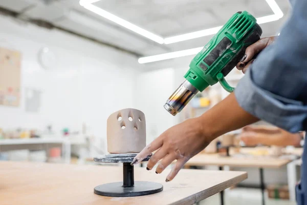 Vue recadrée du maître afro-américain séchant la sculpture en argile avec pistolet à chaleur dans un atelier de poterie — Photo de stock