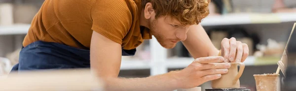 Joven pelirroja artesana en delantal formando taza de arcilla en taller de cerámica borrosa, pancarta — Stock Photo