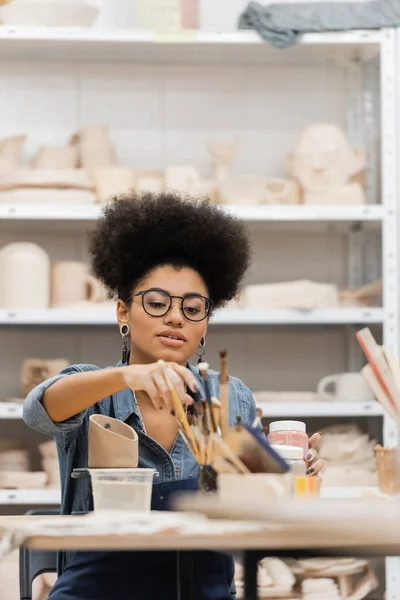 Afroamerikanische Kunsthandwerkerin greift in Töpferwerkstatt zum Pinsel in der Nähe von Tonprodukten — Stockfoto