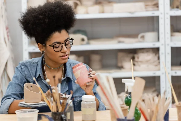 Artisanat afro-américain en lunettes tenant un bocal avec de la peinture près de pinceaux flous et sculpture en argile dans un atelier de poterie — Photo de stock
