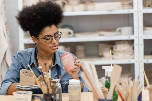 Afroamerikanische Kunsthandwerkerin in Brille hält Glas mit Farbe in der Nähe von verschwommenen Pinseln und Tonskulpturen im Töpferatelier — Stockfoto