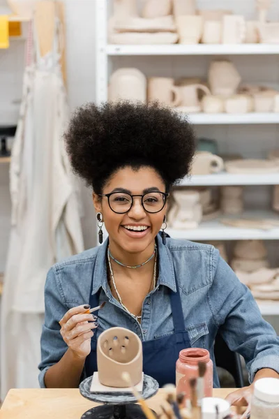 Lächelnde Afroamerikanerin mit Brille hält Pinsel in der Nähe von Tonskulpturen und Farbe in Töpferwerkstatt — Stockfoto