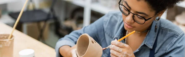 Young african american craftswoman in eyeglasses painting clay product in pottery workshop, banner — Stock Photo