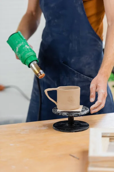 Vue recadrée de l'artisan en tablier tasse d'argile de séchage avec pistolet à chaleur dans l'atelier de poterie — Photo de stock