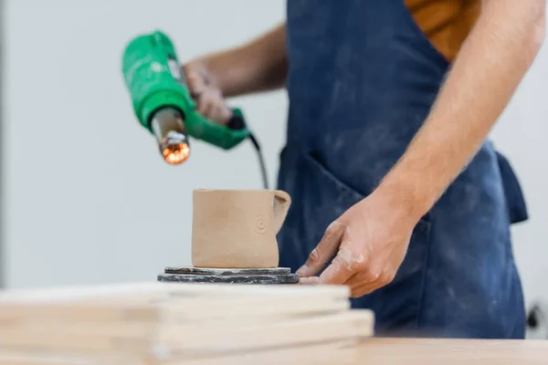 Ausgeschnittene Ansicht von Tonbecher in der Nähe verschwommener Handwerker mit Hitzegewehr im Töpferatelier — Stockfoto