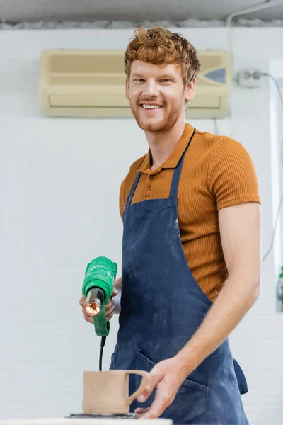 Artesano pelirrojo sonriente en delantal sosteniendo pistola de calor cerca de taza de cerámica borrosa en taller de cerámica - foto de stock
