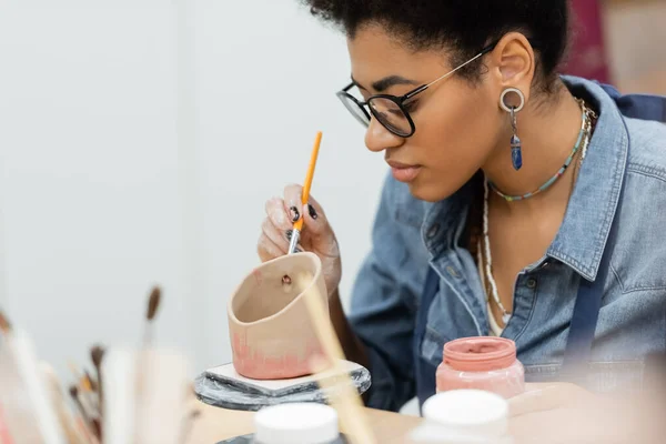 African american artisan in eyeglasses painting on ceramic product near blurred paintbrushes in pottery workshop — Stock Photo