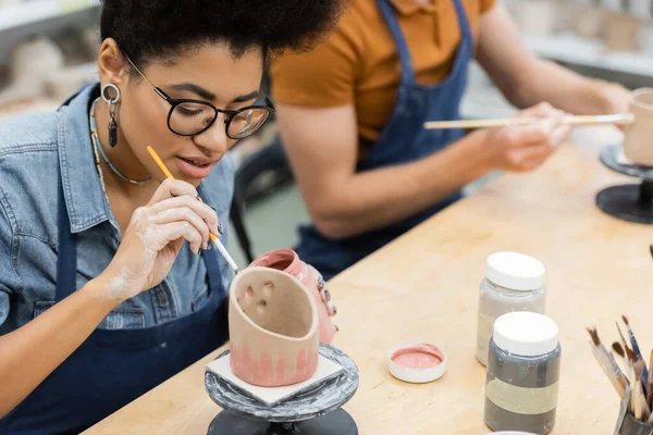 Молода афроамериканська жінка в фартусі на керамічній чашці поруч з розмитим хлопцем під час побачення в гончарній студії — стокове фото