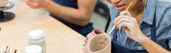 Ausgeschnittene Ansicht einer afrikanisch-amerikanischen Frau in Schürzenmalerei auf Tonerzeugnis in der Nähe eines verschwommenen Freundes im Töpferatelier, Banner — Stockfoto