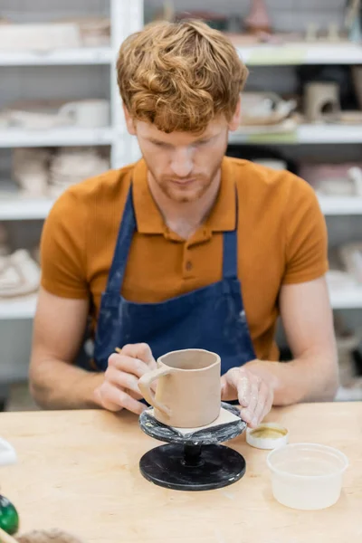 Рыжеволосый ремесленник в фартуке смотрит на керамическую чашку в керамической мастерской — стоковое фото