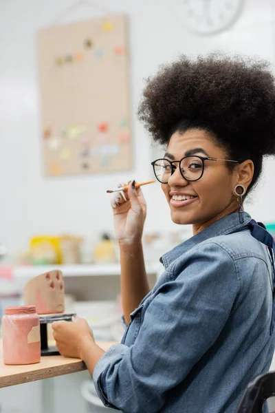 Sorrindo mulher americana africana em óculos segurando pincel perto de escultura de argila e pintura em estúdio de cerâmica — Fotografia de Stock