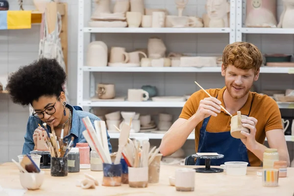 Couple multiethnique positif peignant des sculptures en argile pendant la date en atelier de poterie — Photo de stock
