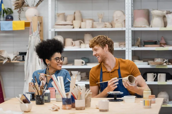 Homme souriant regardant petite amie afro-américaine près de l'argile et pinceaux en atelier de poterie — Photo de stock