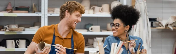 Smiling redhead man painting on clay product near african american girlfriend in pottery workshop, banner — Stock Photo