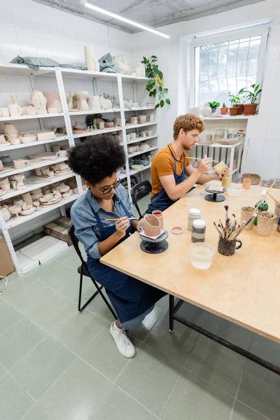 Pareja multiétnica en delantales pintando sobre productos cerámicos en estudio de cerámica - foto de stock