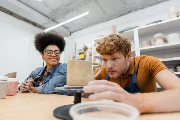 Redhead man looking at clay cup near smiling african american girlfriend in pottery studio — Stock Photo