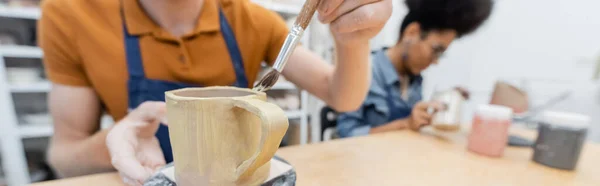 Hombre borroso pintando en taza de cerámica cerca de novia afroamericana en taller de cerámica, pancarta - foto de stock