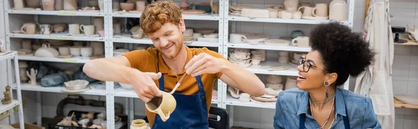 Redhead man painting on clay cup near cheerful african american girlfriend in pottery workshop, banner — Stock Photo