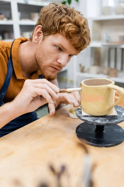 Jeune artisan rousse peignant sur une tasse d'argile dans un atelier de poterie — Photo de stock