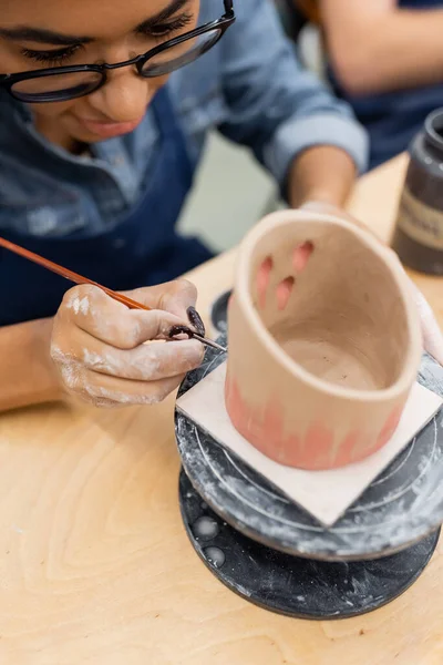 Jovem afro-americana em óculos pintando em produto de barro desfocado no estúdio de cerâmica — Fotografia de Stock