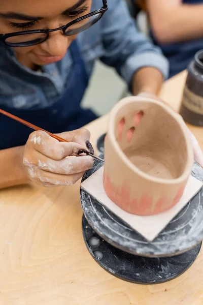 Young african american woman in eyeglasses painting on blurred clay product in pottery studio — Stock Photo