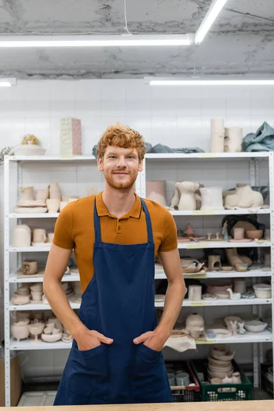 Smiling redhead artisan in apron looking at camera in pottery studio — Stock Photo