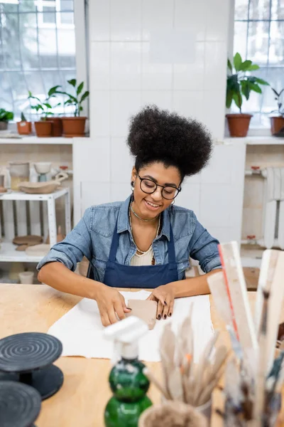 Улыбающаяся американка в фартуке держит глину на столе в мастерской по гончарному делу — стоковое фото