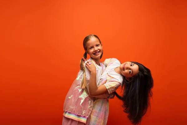 Веселая девушка обнимает подругу брюнетку, улыбаясь в камеру, изолированную от оранжевого — стоковое фото