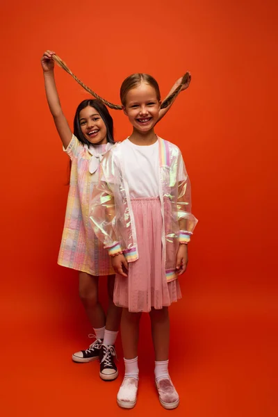 Full length of excited girl holding pigtails of cheerful friend on orange background — Stock Photo