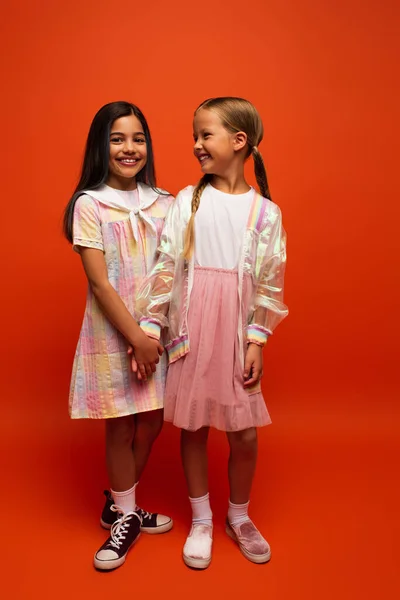 Full length of brunette girl in dress holding hands with friend in rain jacket on orange background — Stock Photo