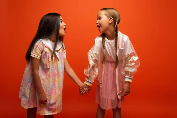 Preteen girls with open mouths holding hands and looking at each other while having fun isolated on orange — Stock Photo