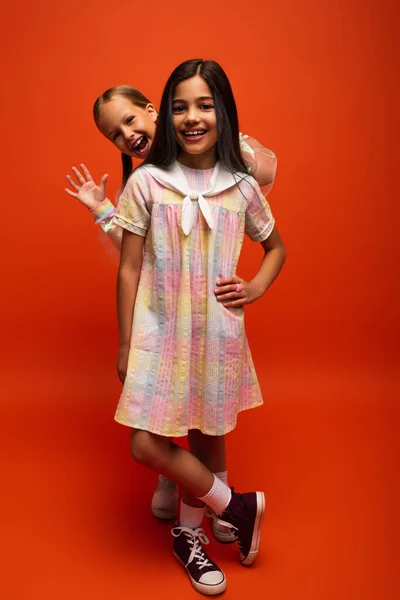 Excitada chica agitando la mano detrás de amigo en vestido posando sobre fondo naranja - foto de stock