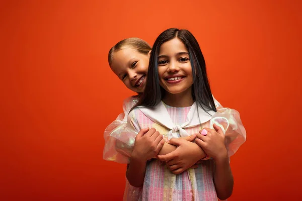 Збуджена дівчина обіймає подругу брюнетки, дивлячись на камеру ізольовано на помаранчевому — стокове фото