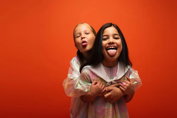 Amigos emocionados abrazando y sobresaliendo lenguas mientras se divierten aislados en naranja - foto de stock