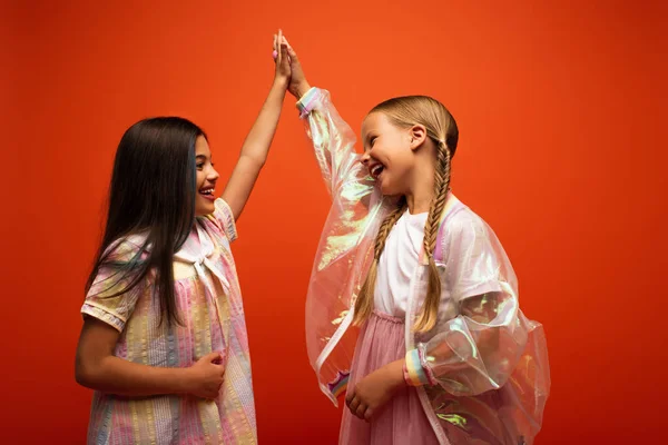 Happy girls in dress and transparent rain jacket giving high five isolated on orange — Stock Photo