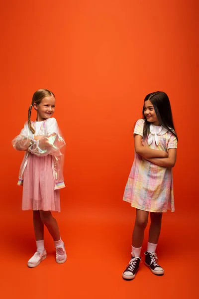 Longitud completa de niñas preadolescentes alegres de pie con los brazos cruzados y sonriendo el uno al otro sobre fondo naranja - foto de stock