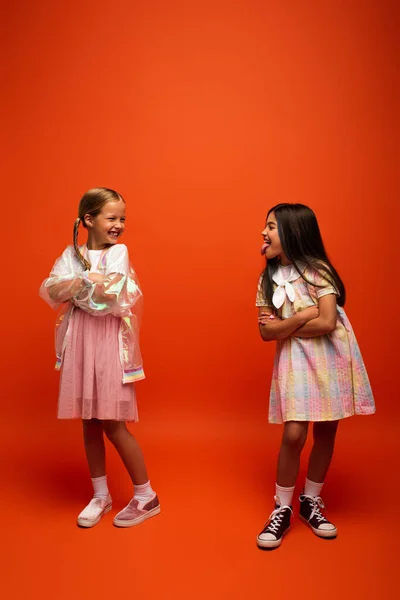 Volle Länge der brünetten Mädchen streckt Zunge zu lächelnden Freund stehend mit verschränkten Armen auf orangefarbenem Hintergrund — Stockfoto
