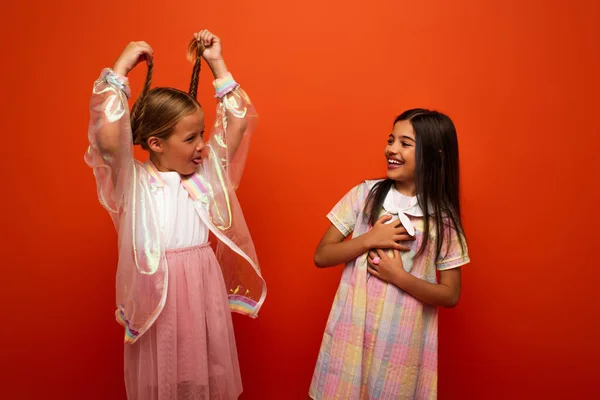 Brunette girl touching chest and laughing near friend holding her pigtails and sticking out tongue isolated on orange — Stock Photo