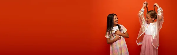 Funny girl holding pigtails and sticking out tongue near laughing friend isolated on orange, banner — Stock Photo