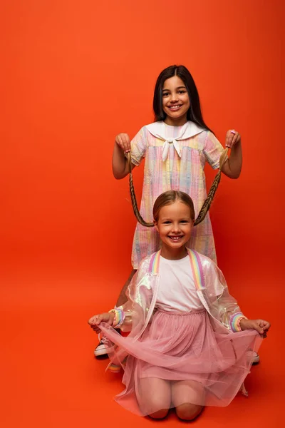 Smiling brunette girl holding pigtails of cheerful friend sitting on orange background — Stock Photo