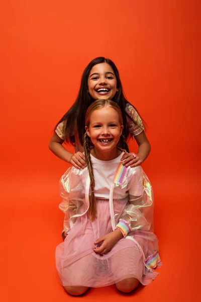 Alegre chica en chaqueta de lluvia sonriendo a la cámara mientras está sentado cerca emocionado amigo sobre fondo naranja - foto de stock