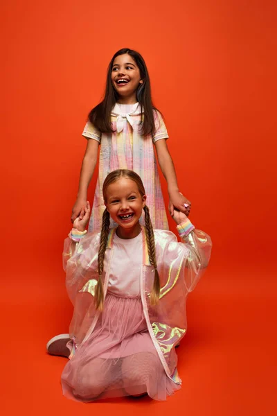 Feliz morena chica cogida de la mano con alegre amigo sentado en naranja fondo - foto de stock