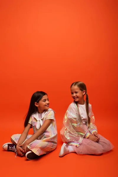Joyeuses filles en robe et veste de pluie assis et souriant à l'autre sur fond orange — Photo de stock