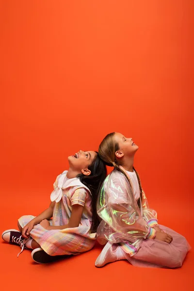 Menina surpreso olhando para cima enquanto sentado de volta para trás com o amigo sorrindo com olhos fechados no fundo laranja — Fotografia de Stock