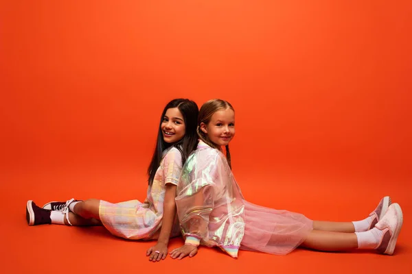 Longitud completa de las niñas con estilo en la ropa de moda sentado espalda con espalda y mirando a la cámara en fondo naranja - foto de stock