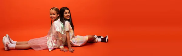 Full length of happy and trendy girls sitting back to back and looking at camera on orange background, banner — Stock Photo