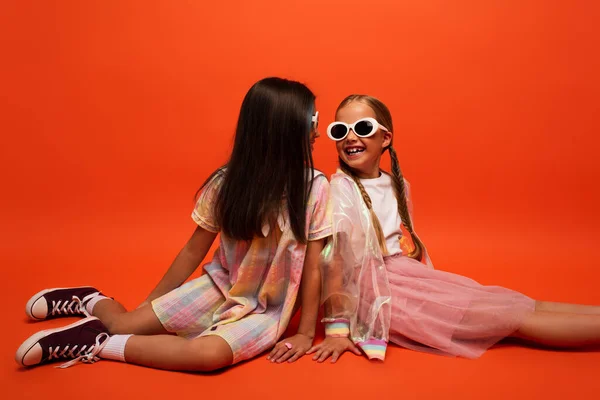 Élégant preteen enfants en lunettes de soleil souriant à l'autre tout en étant assis sur fond orange — Photo de stock